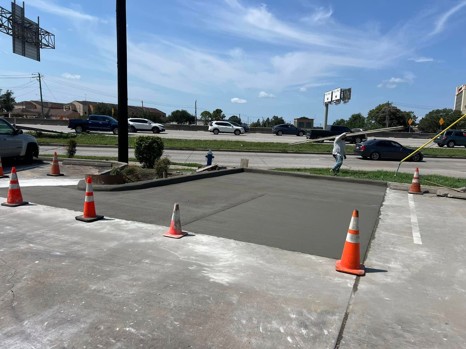 Newly poured concrete patch surrounded by orange cones in a parking area, with a person walking nearby.
