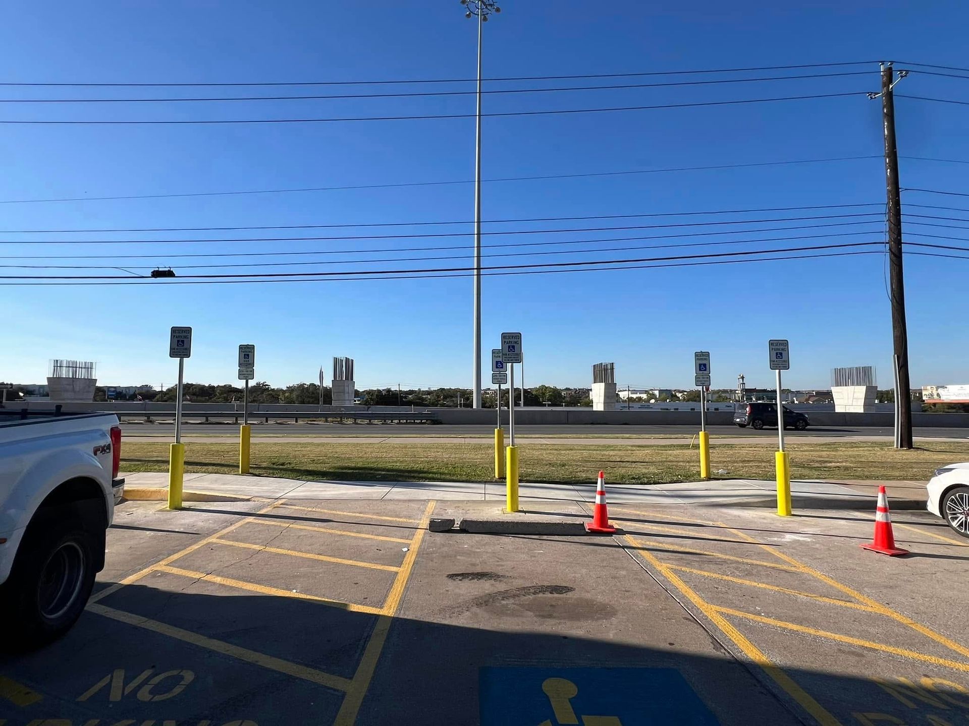 Parking area with signs, bollards, and cones. Buildings in the distance. Blue sky overhead.