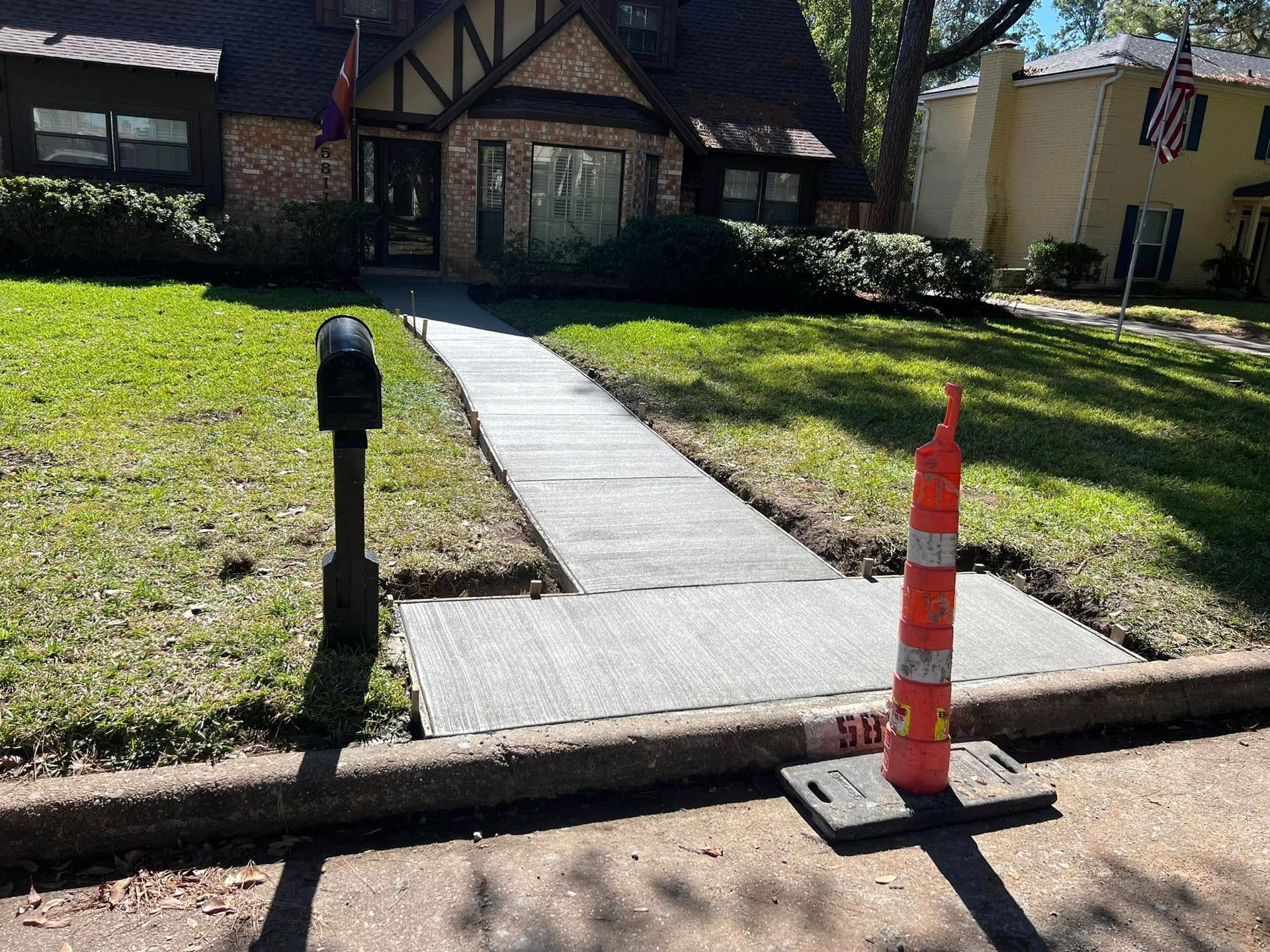 Newly poured concrete sidewalk leading to a Tudor style house; an orange traffic cone sits at the sidewalk's edge.