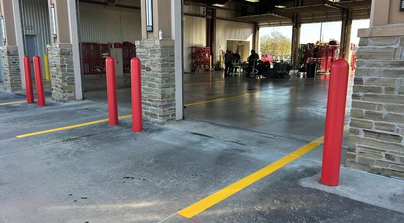 Exterior view of a fire station with red bollards and yellow lines. People and fire trucks inside the open bay.