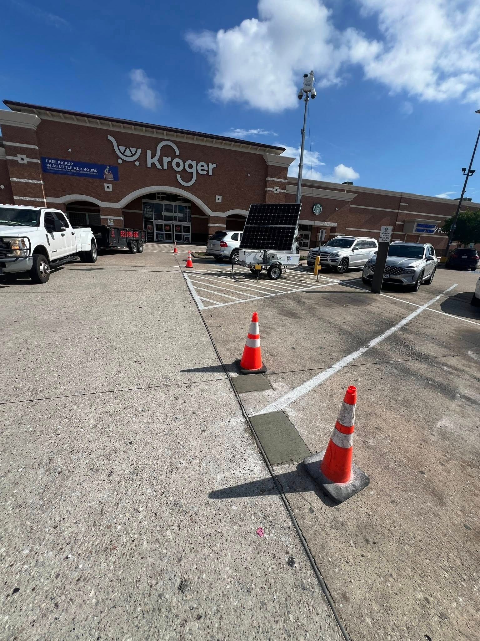 Kroger store exterior with vehicles, orange cones, and a solar-powered tower on a sunny day.