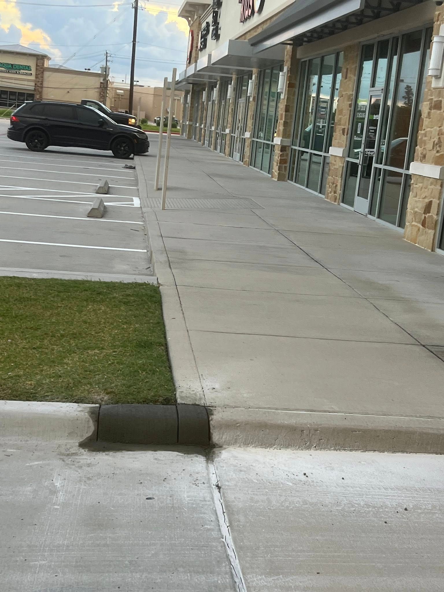 Sidewalk with curb, leading to a row of storefronts. Black car parked in lot. Green grass. Cloudy sky.