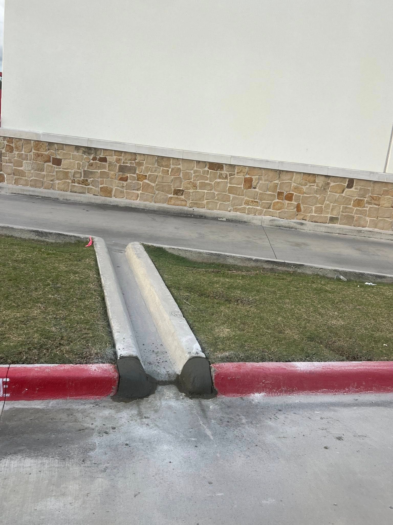 Concrete drainage channel on a slight incline, red curb, grass, and a stone and white building in the background.