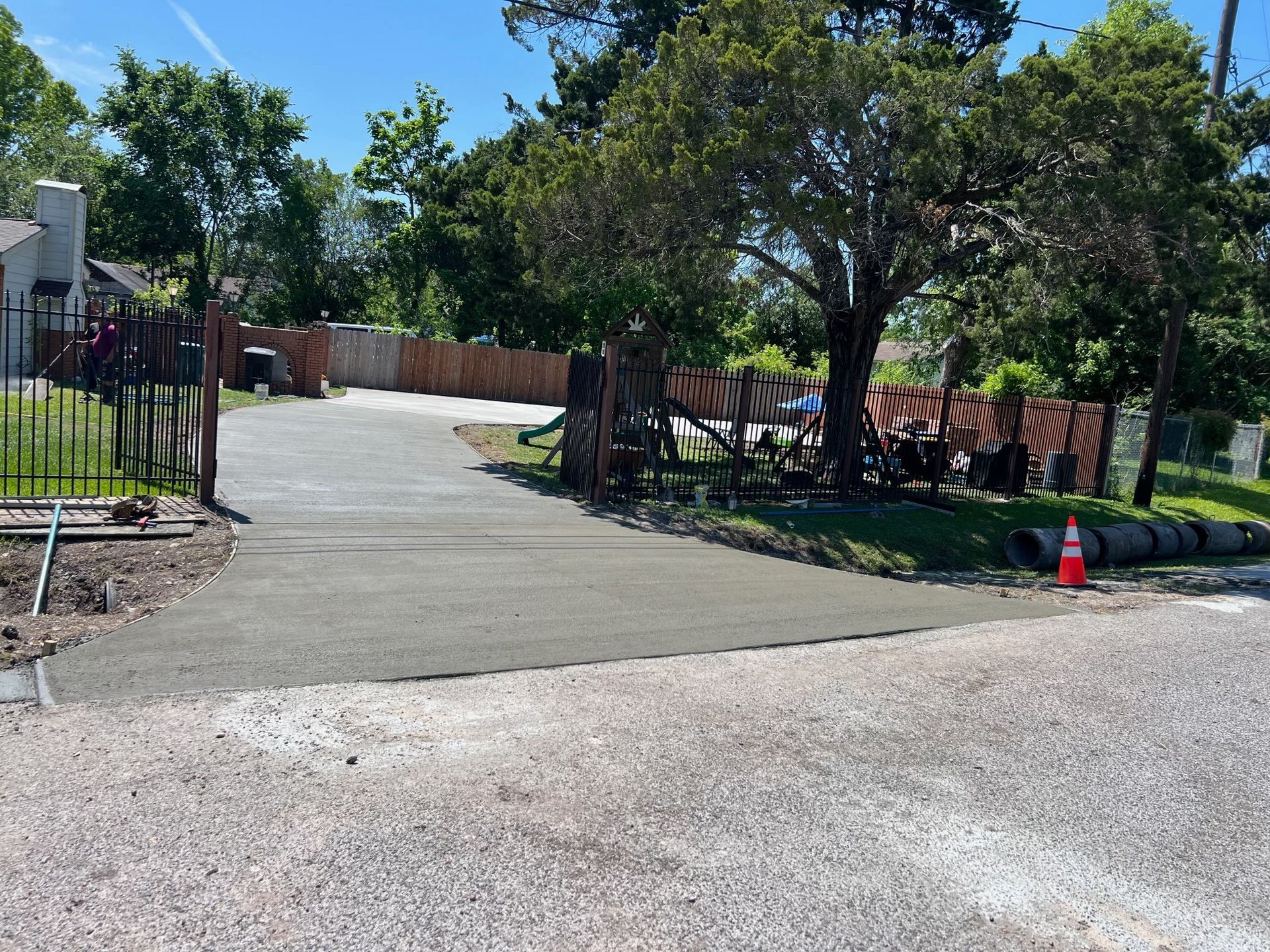 Newly poured concrete driveway leading to a house, gravel road in the foreground, trees and fencing in background.