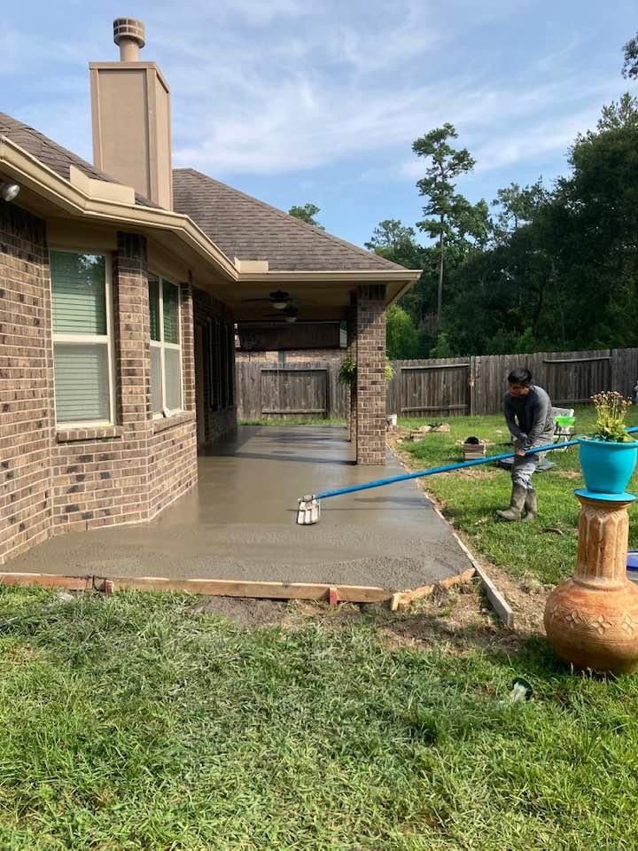 Man leveling wet concrete on a patio with a long-handled tool, next to a brick house and grass.