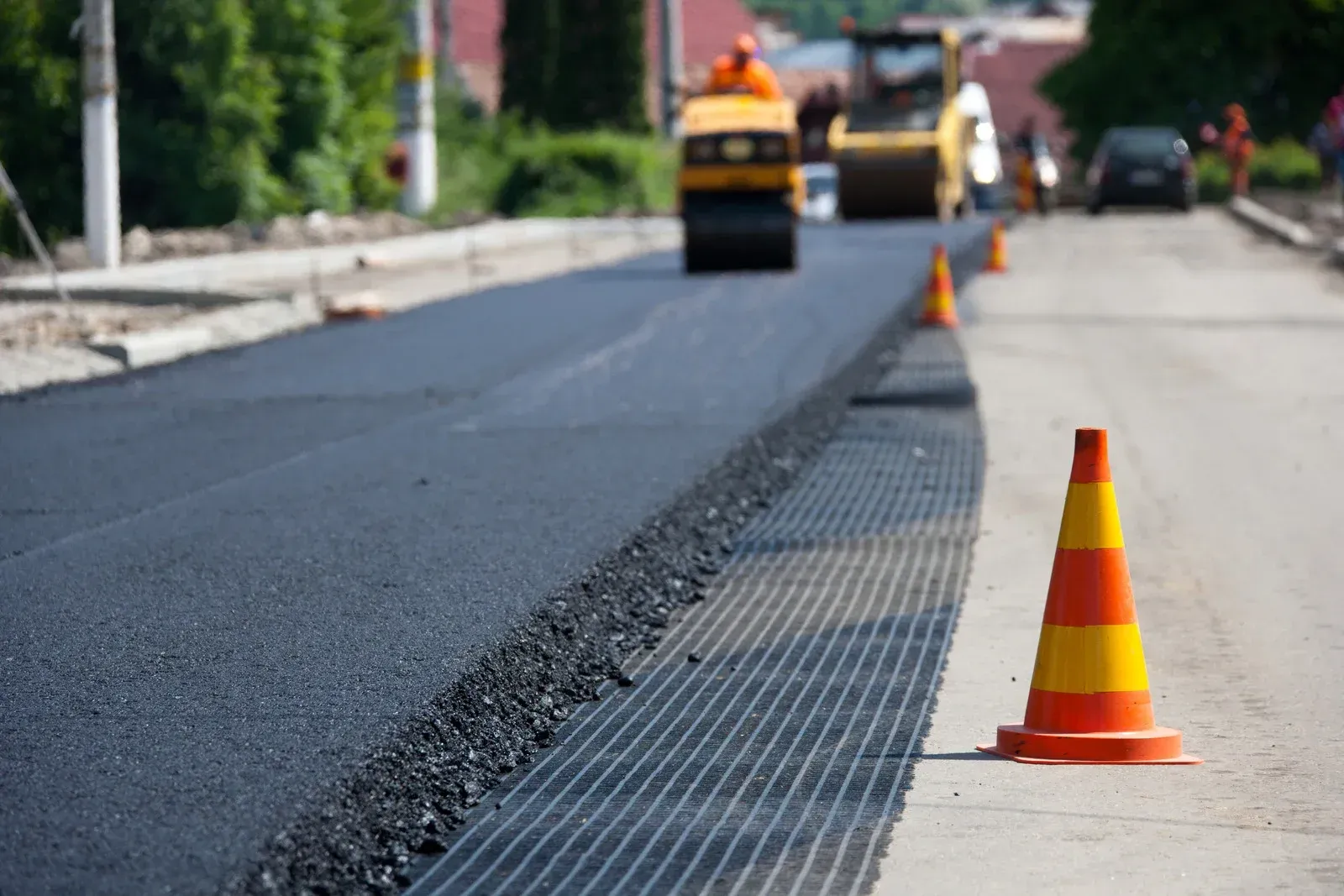 Road resurfacing: Fresh asphalt next to a safety cone, with construction equipment and workers in the blurred background.