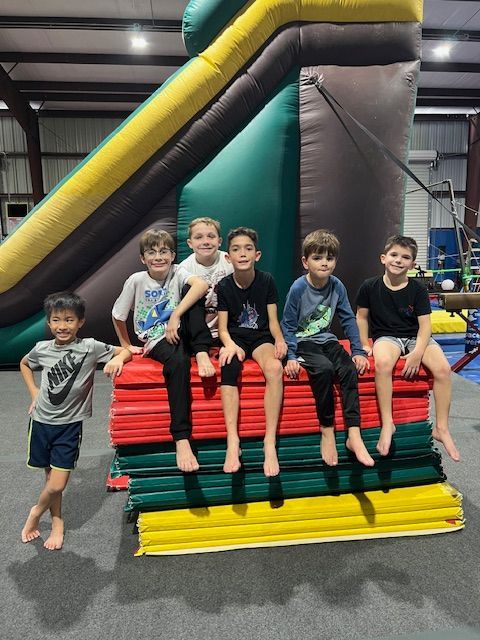 A group of young boys are sitting on a stack of mats in front of an inflatable slide.