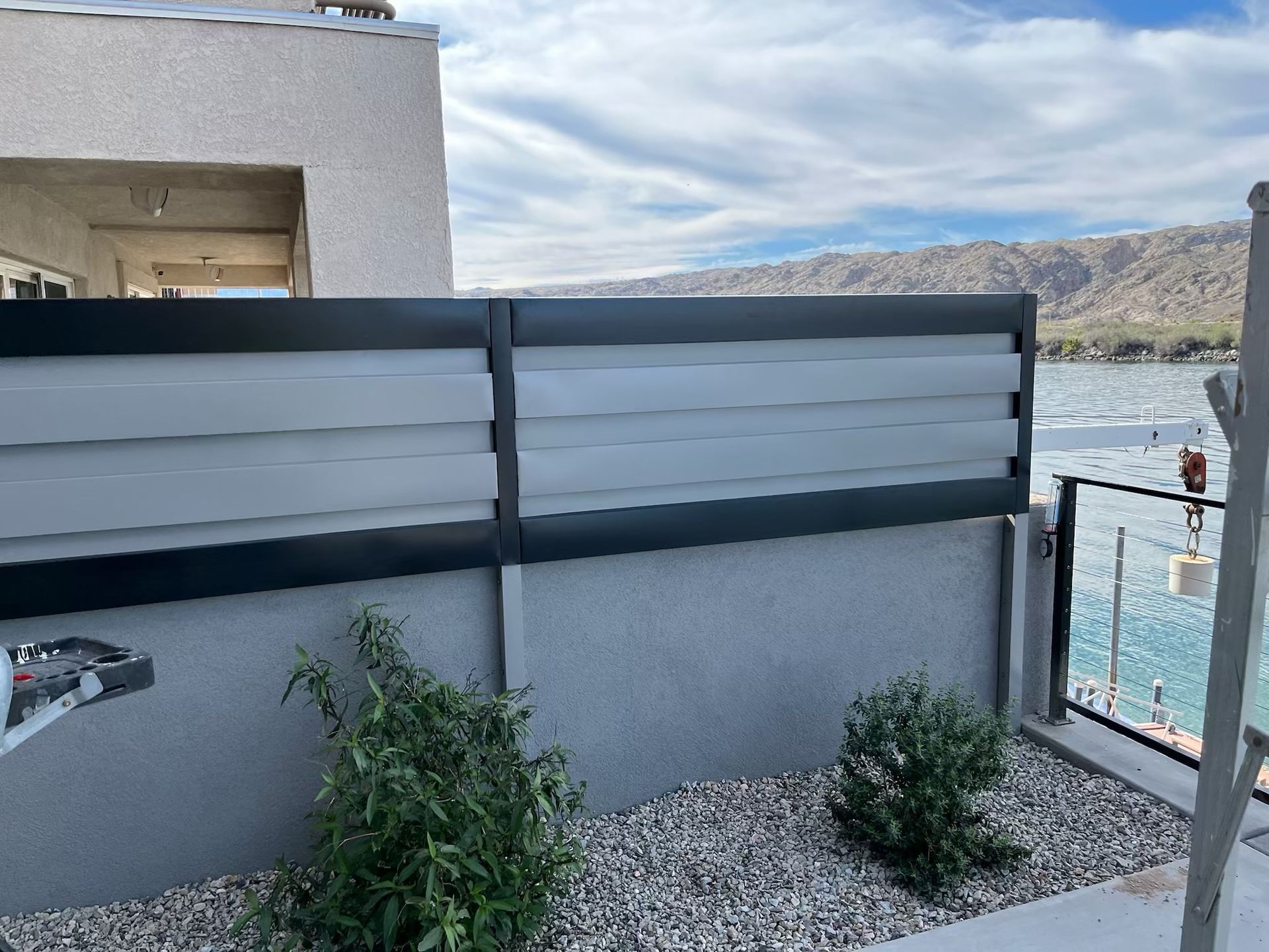 A black-framed privacy fence with grey horizontal slats overlooks a lake with mountains in the background.