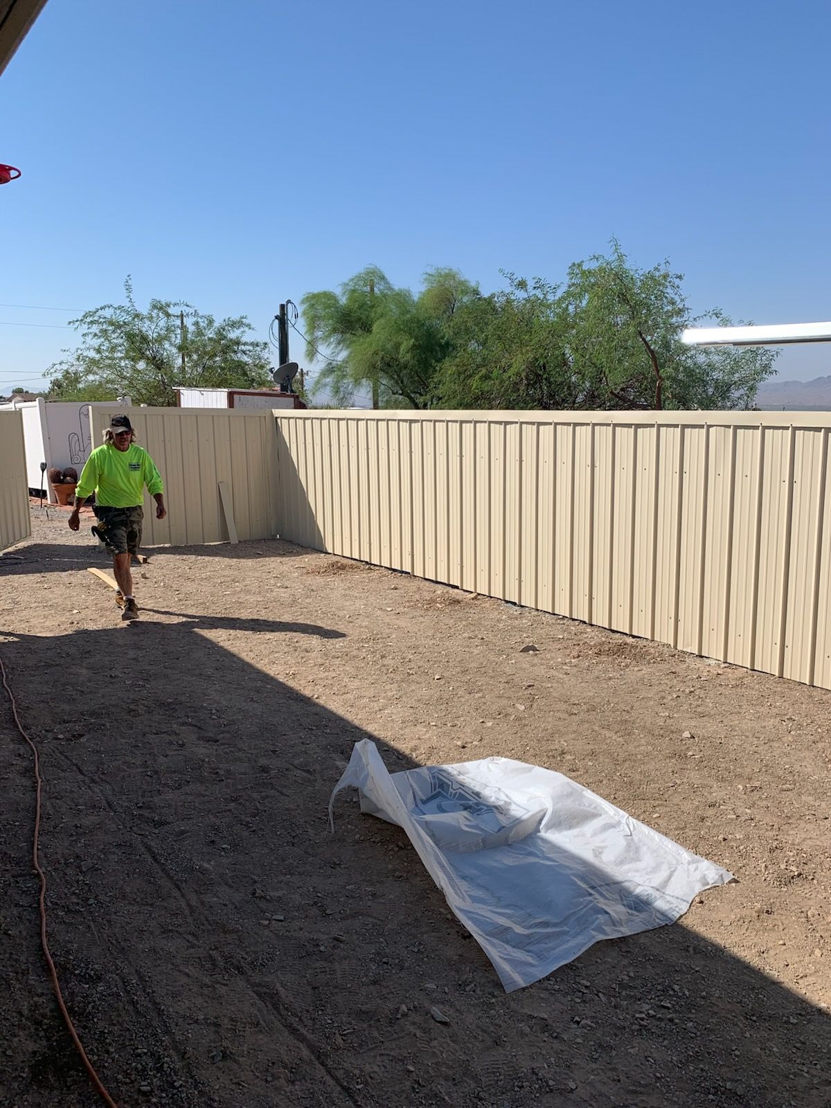 A person in a neon yellow shirt walks across a gravel yard near a tan metal fence under a clear blue sky.