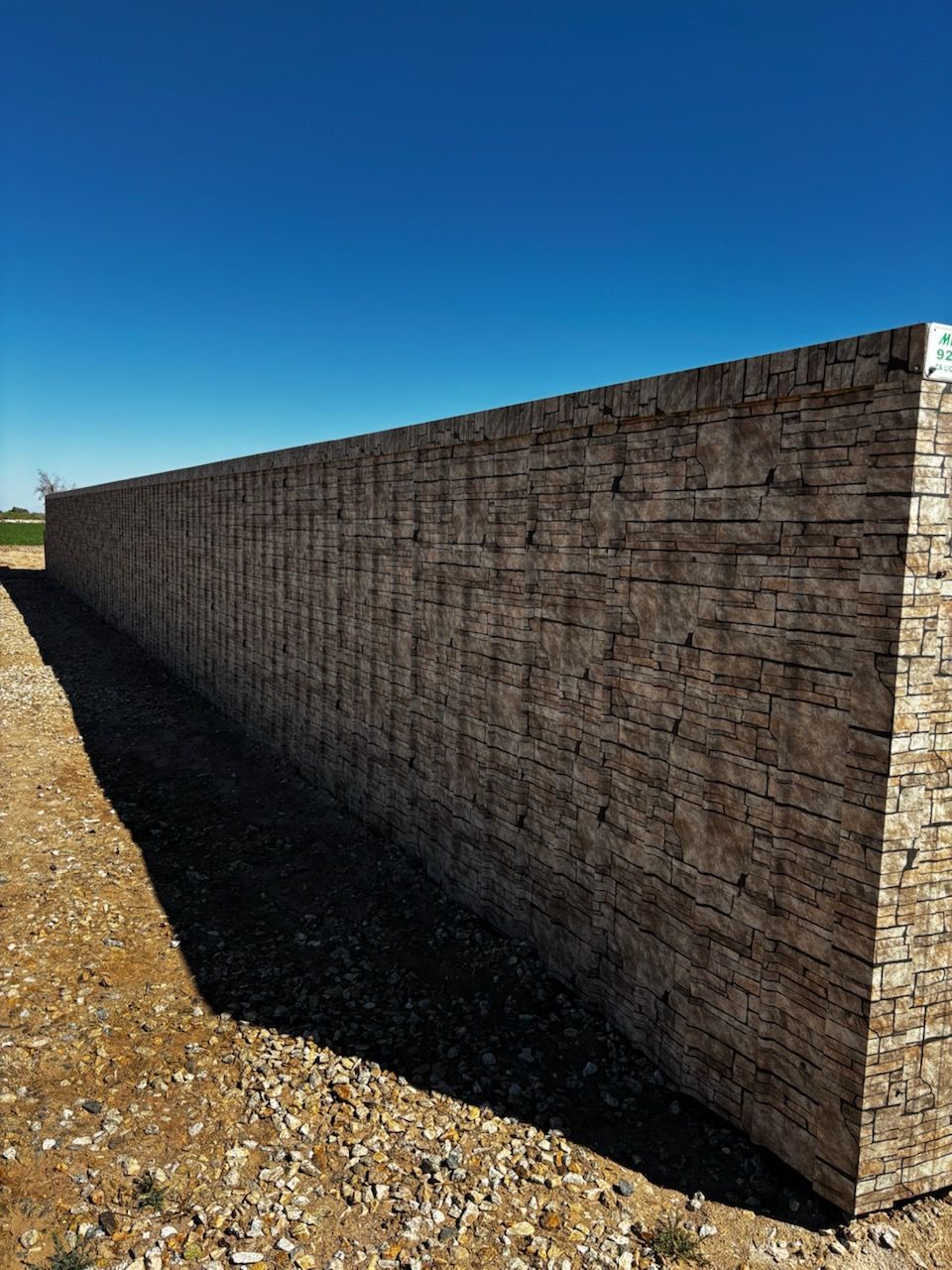 A long, textured stone retaining wall stretches across a gravel field under a clear blue sky.