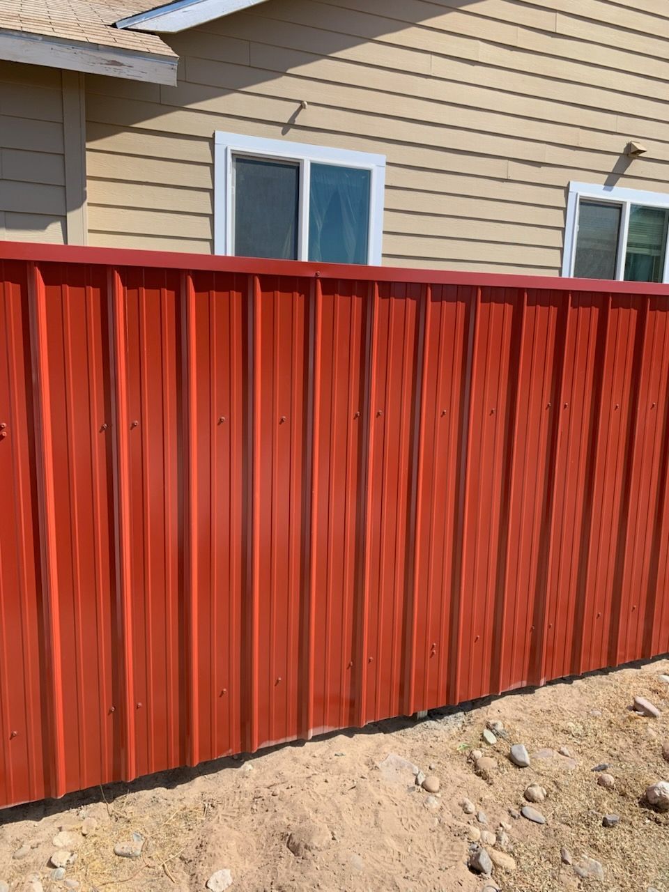 A red metal fence stands in front of a tan house with two white-framed windows on a sunny day.