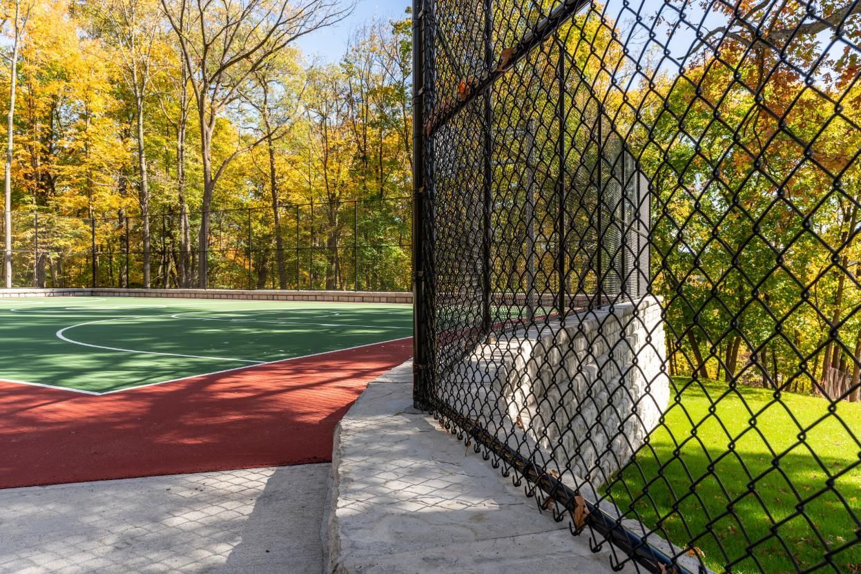 A black chain-link fence surrounds a sports court with a red and green surface, trees in background.