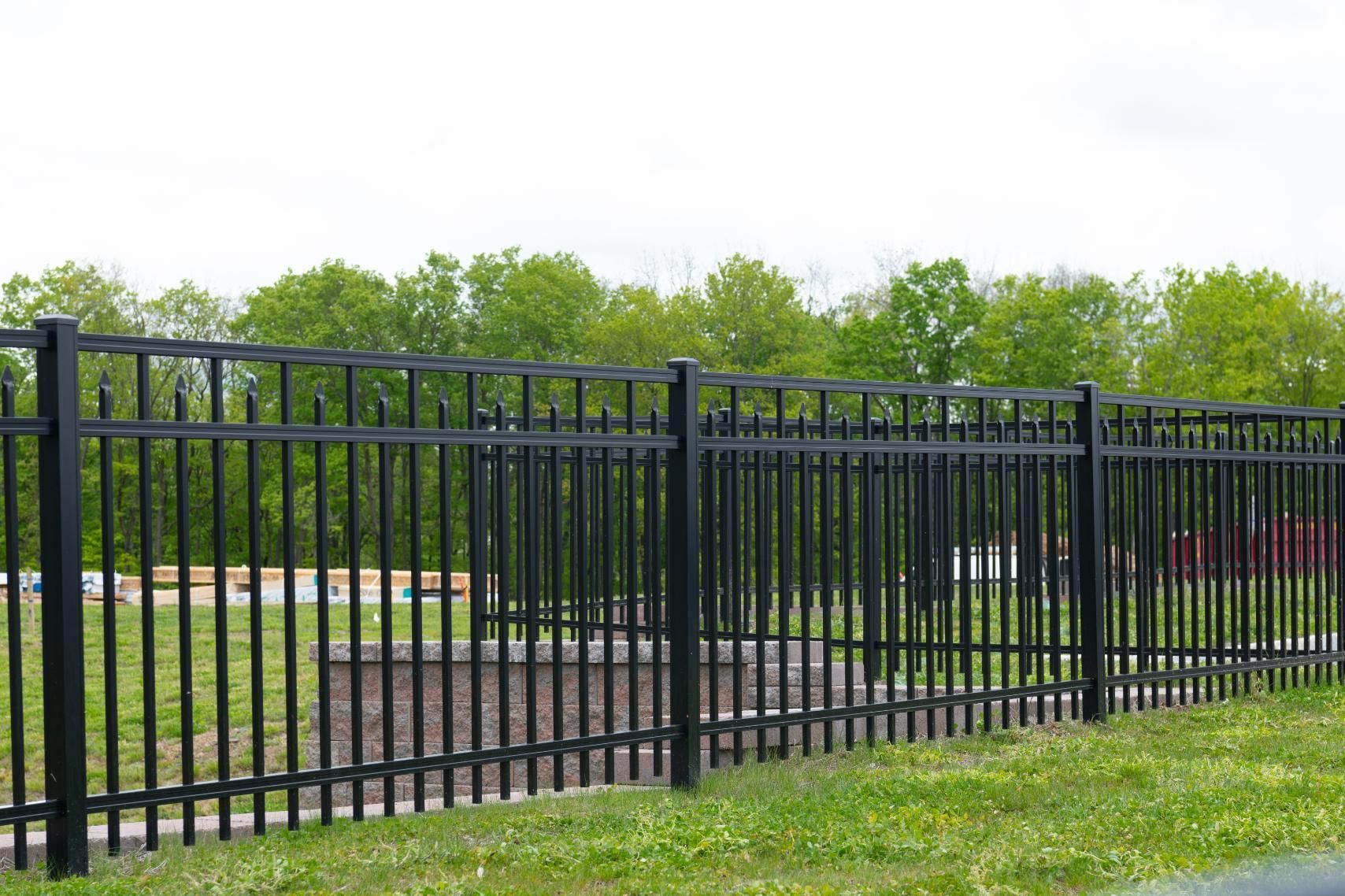 Black metal fence bordering a grassy area with trees in the background under a cloudy sky.