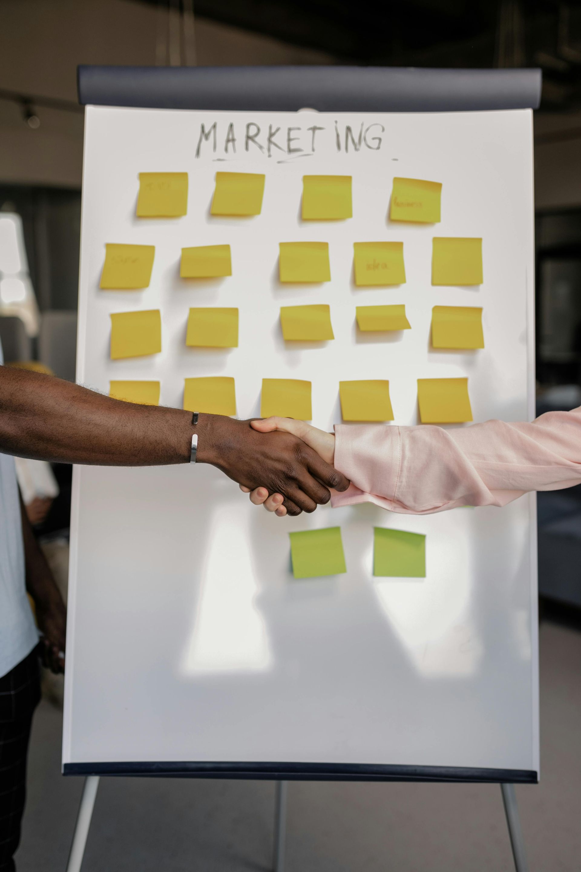 Two people are shaking hands in front of a whiteboard with sticky notes on it.