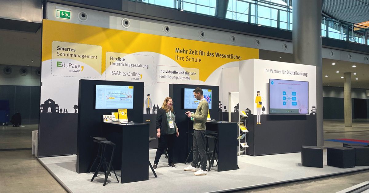 Großer Messestand in RAABE-Corporate-Design, etwa 3 Meter hoch mit einer großflächigen Wand mit Illustrationen und Info-Blasen. Davor stehen zwei Tische mit Monitoren und ein Quadrat mit einem größeren Monitor. Oben drüber hängt ein großer Würfel mit dem RAABE-Logo.