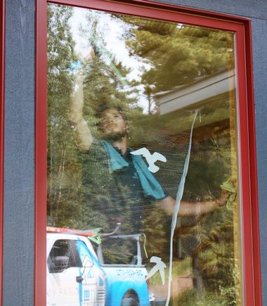 Person seen through a red-framed window, reflected in glass beside trees and a white-blue vehicle.