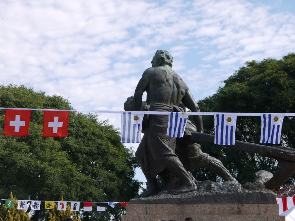 Estatua de dos hombres tirando de un bote, ondeando banderas suizas y uruguayas contra un cielo nublado.