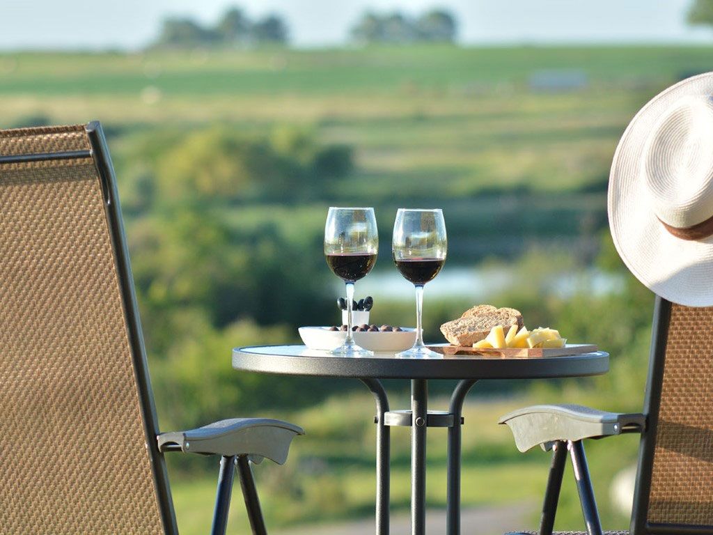 Mesa puesta para dos con copas de vino, bocadillos y un sombrero; con vista a un paisaje verde borroso.