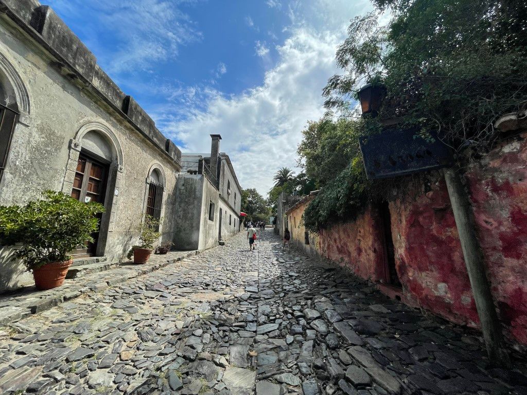 Calle adoquinada en Colonia del Sacramento, Uruguay. Edificios históricos bordean la calle bajo un cielo parcialmente nublado.