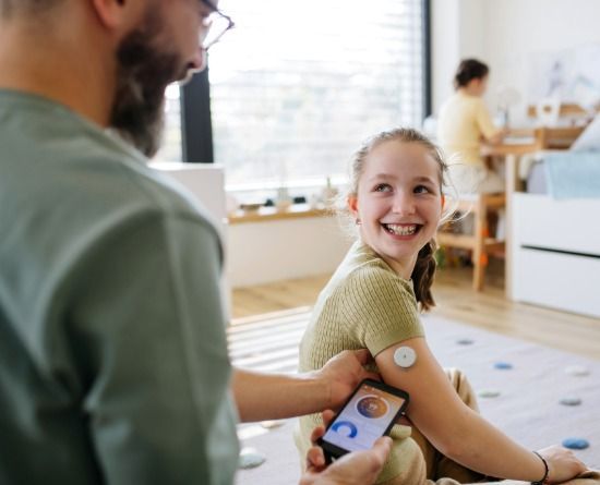 Man checks glucose monitor on girl's arm; both smile indoors.