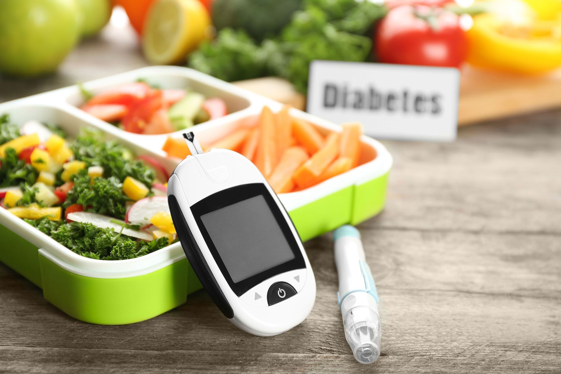A diabetic test kit and a pen are on a wooden table next to a lunch box filled with vegetables.