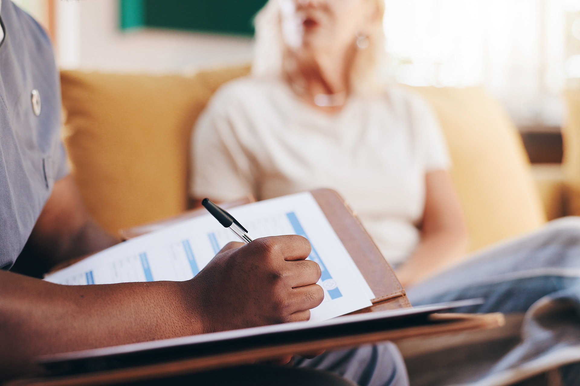 Person writing on a clipboard during a counseling session, with a blurred seated patient in the background