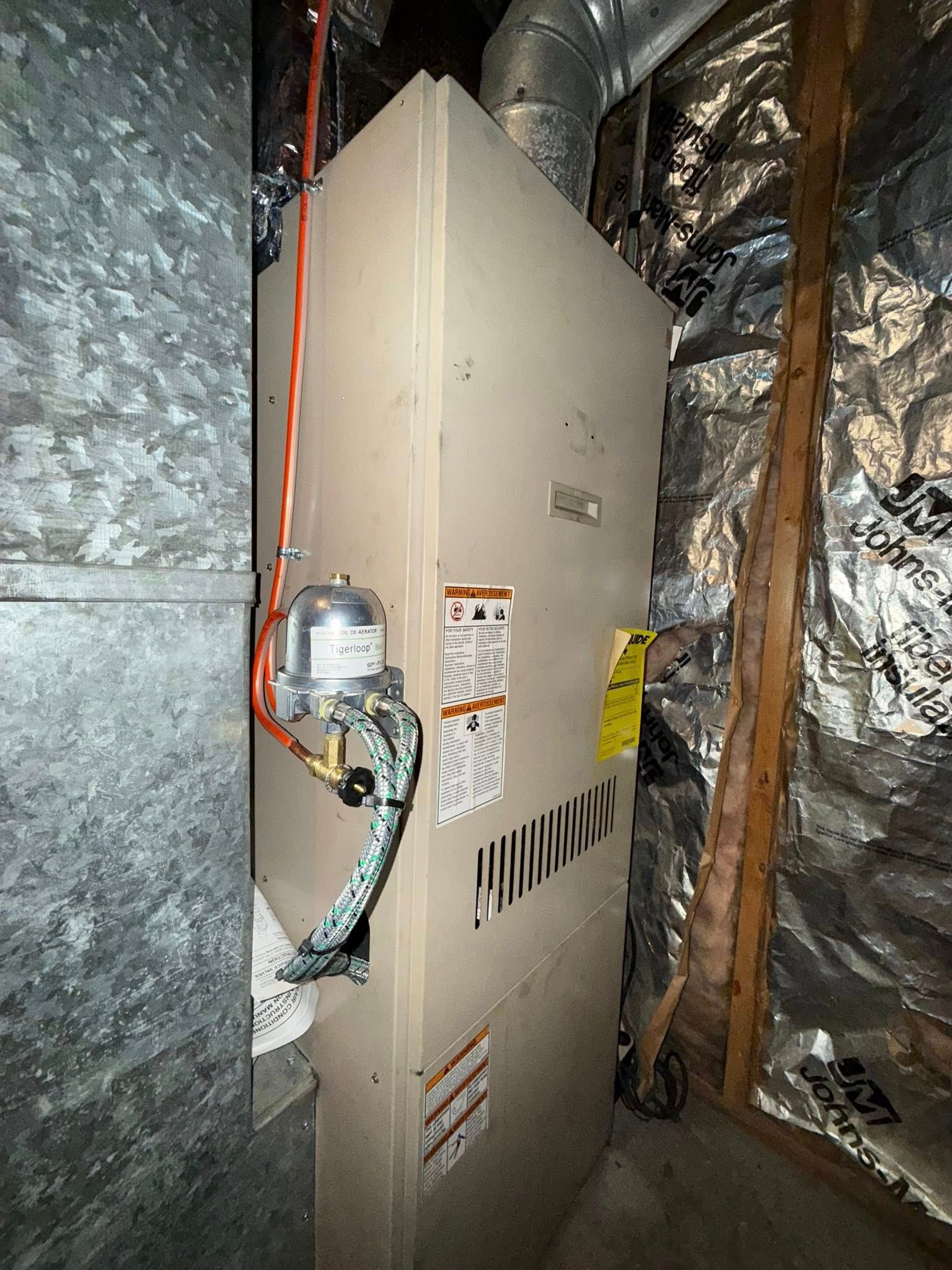 Furnace unit in an enclosed space with ductwork and insulation. Beige unit, various warning labels.