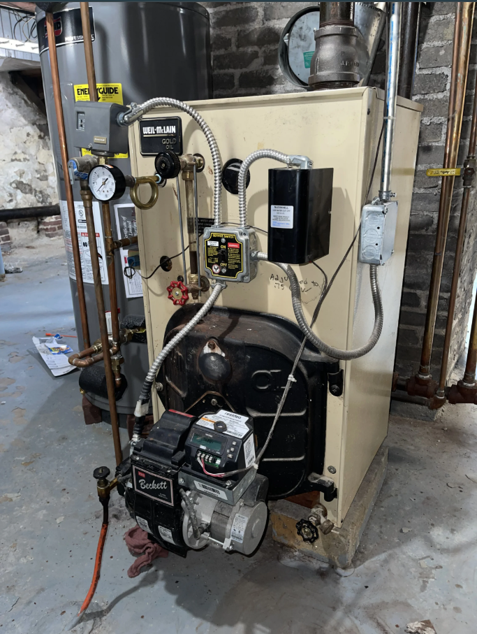 Oil-fired boiler in a basement setting, with various gauges, pipes, and electrical components visible.