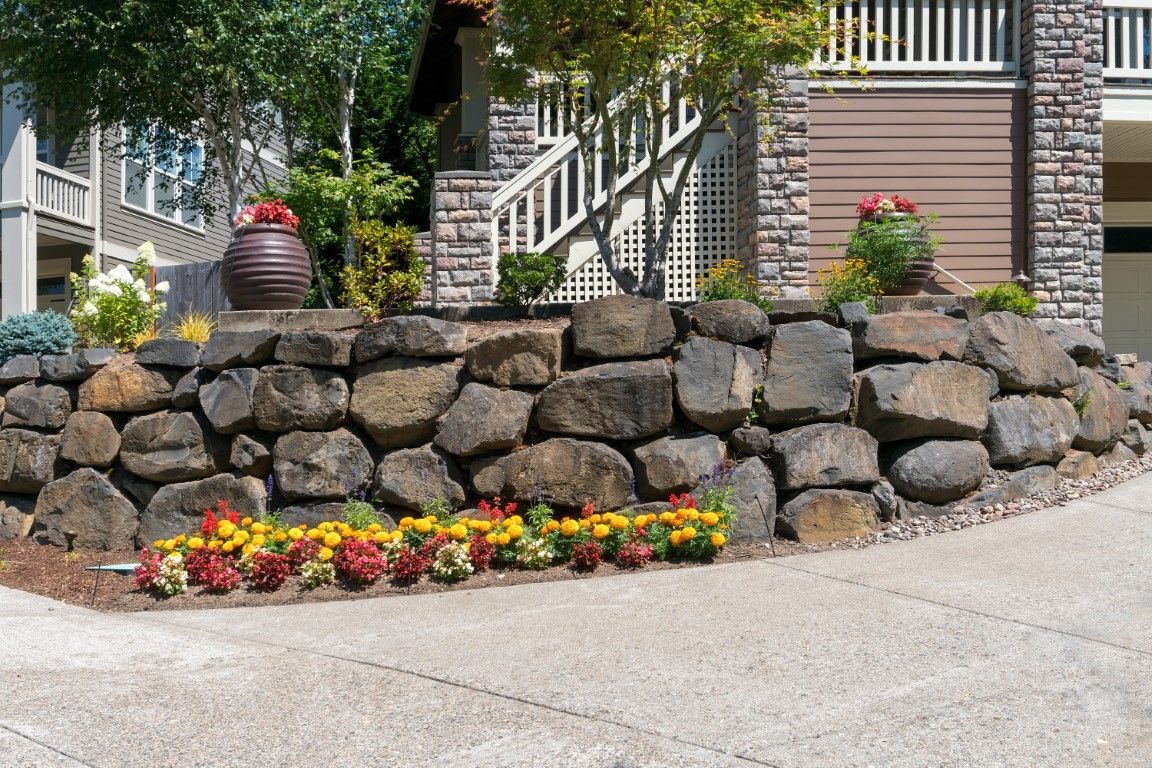 Stone retaining wall with colorful flowers and decorative urns, in front of a building with a stairway.