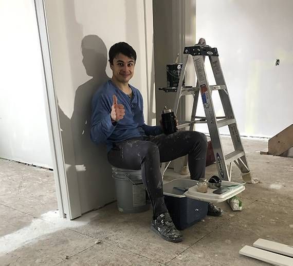 Alex is sitting on a bucket next to a ladder and giving a thumbs up.
