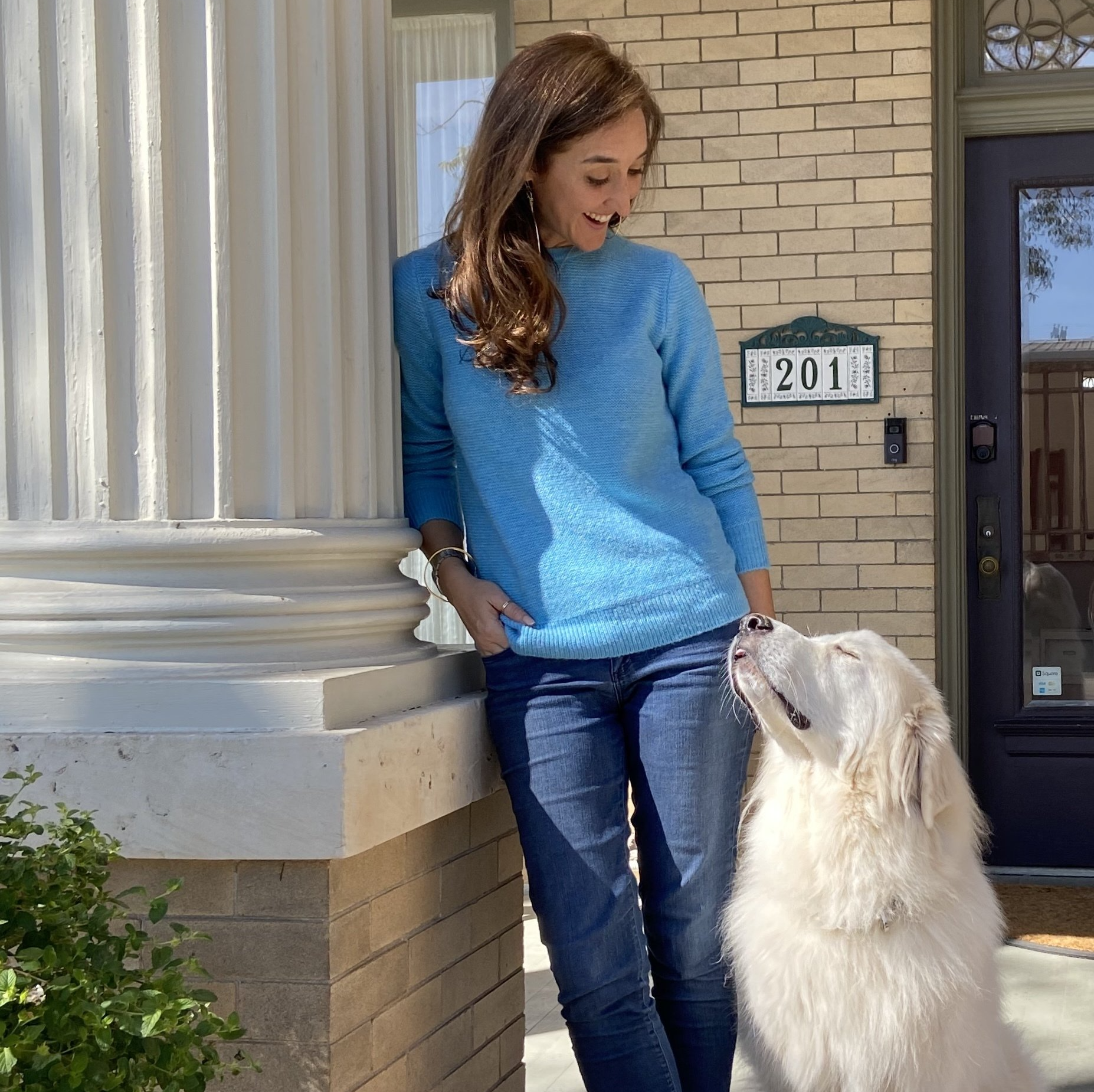 A woman in a blue sweater is standing next to a white dog on a porch.