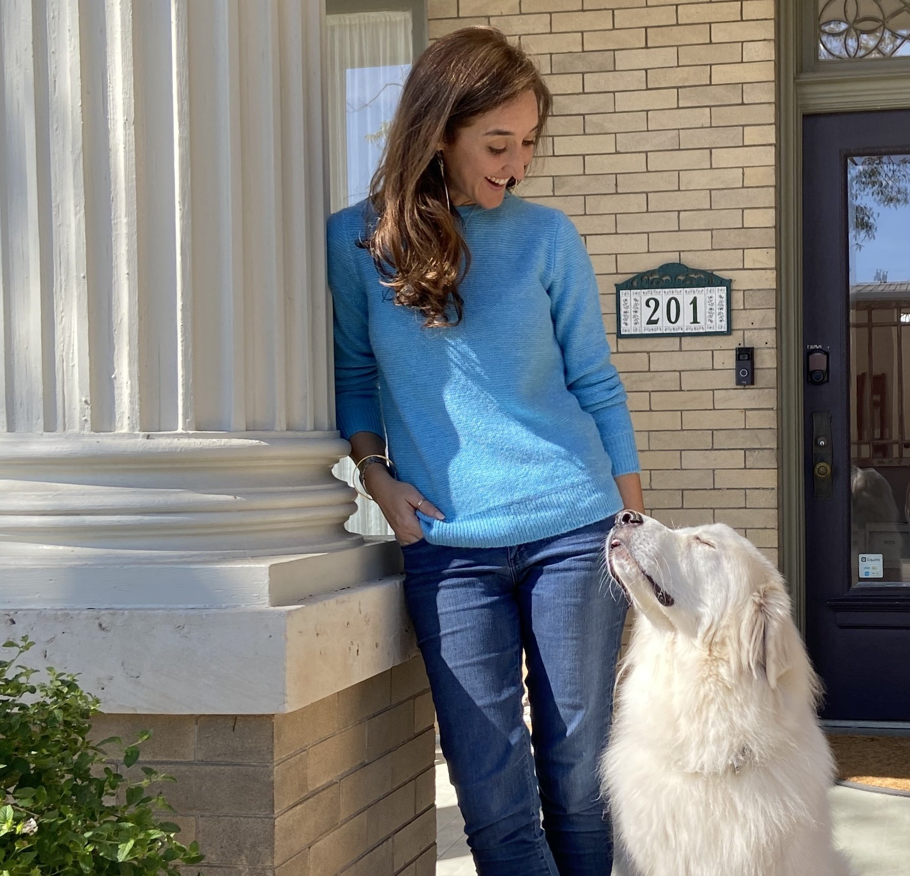A woman in a blue sweater is standing next to a white dog on a porch
