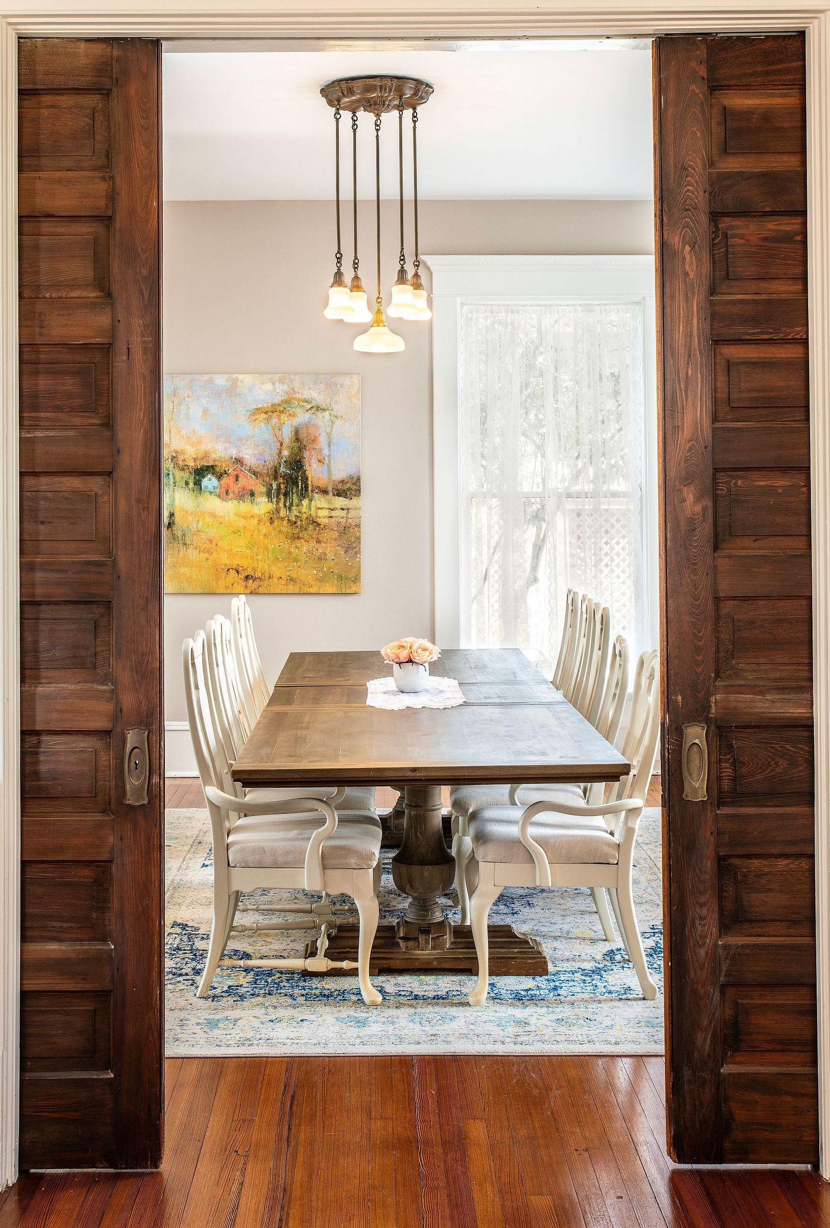 A dining room with a wooden table and chairs and a painting on the wall.