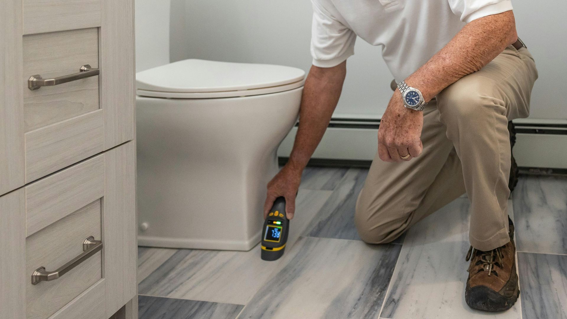 A professional inspects the base of a bathroom toilet using a handheld moisture meter on a patterned floor.