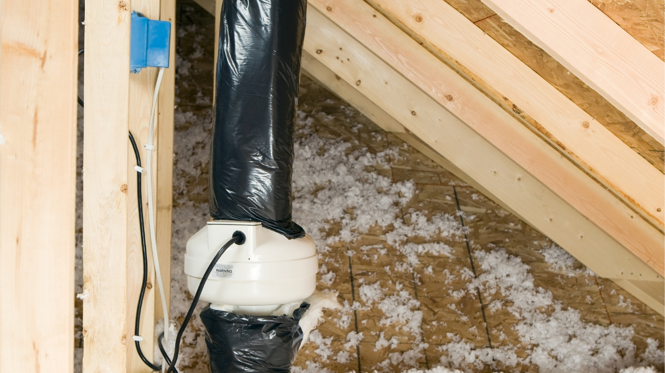 Radon mitigation fan installed in an attic with black piping and an electrical box mounted to a wooden stud.