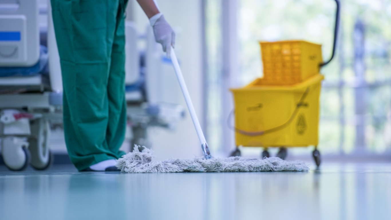 A person in green scrubs mopping a hospital floor near a patient bed, with a yellow cleaning cart in the background.