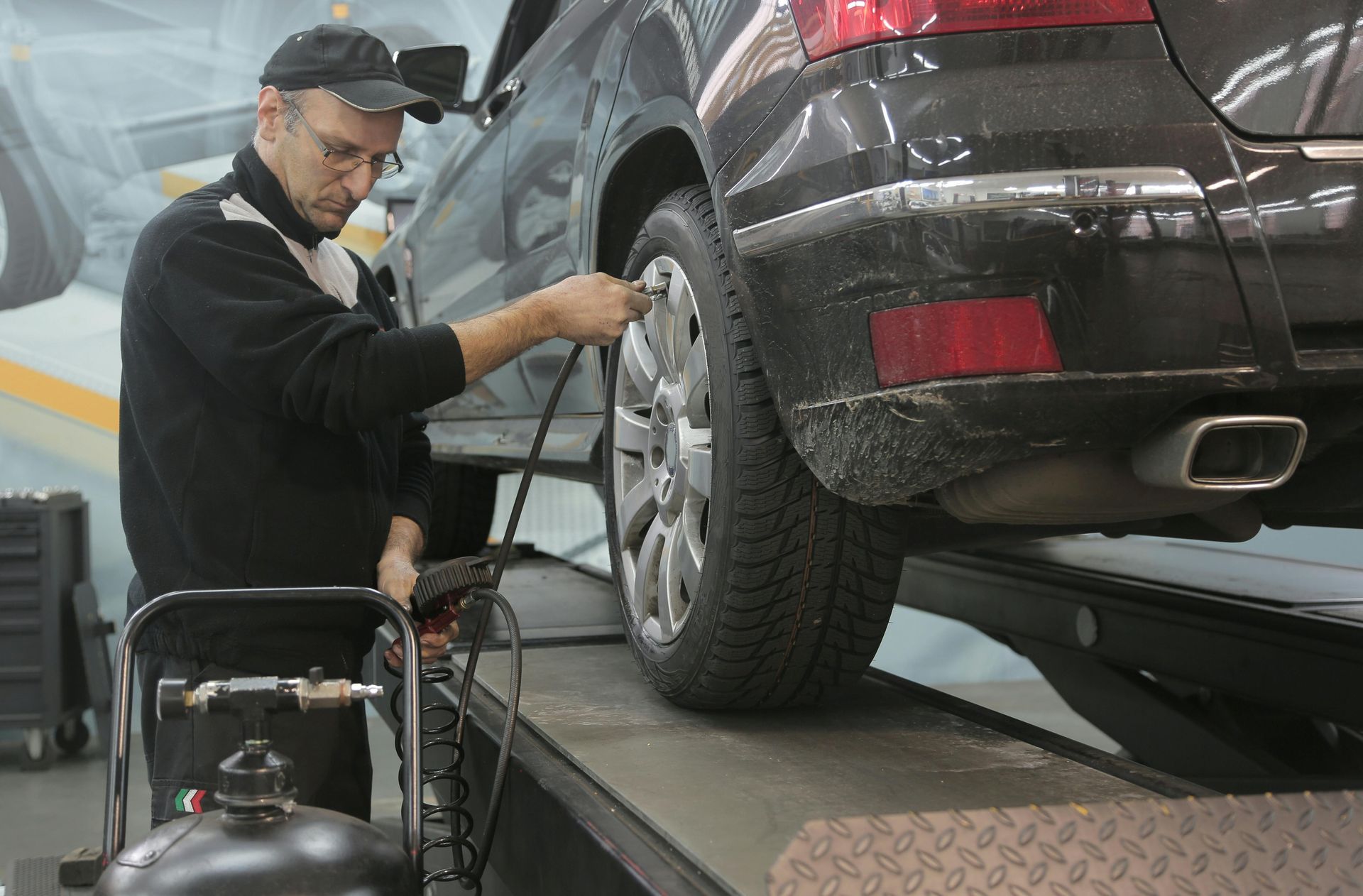 Mechanic inflating tire on a black car at a garage. He wears a cap, holding air pump.
