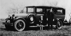Vintage hearse parked outdoors with two people, one adult and one child, standing next to it.