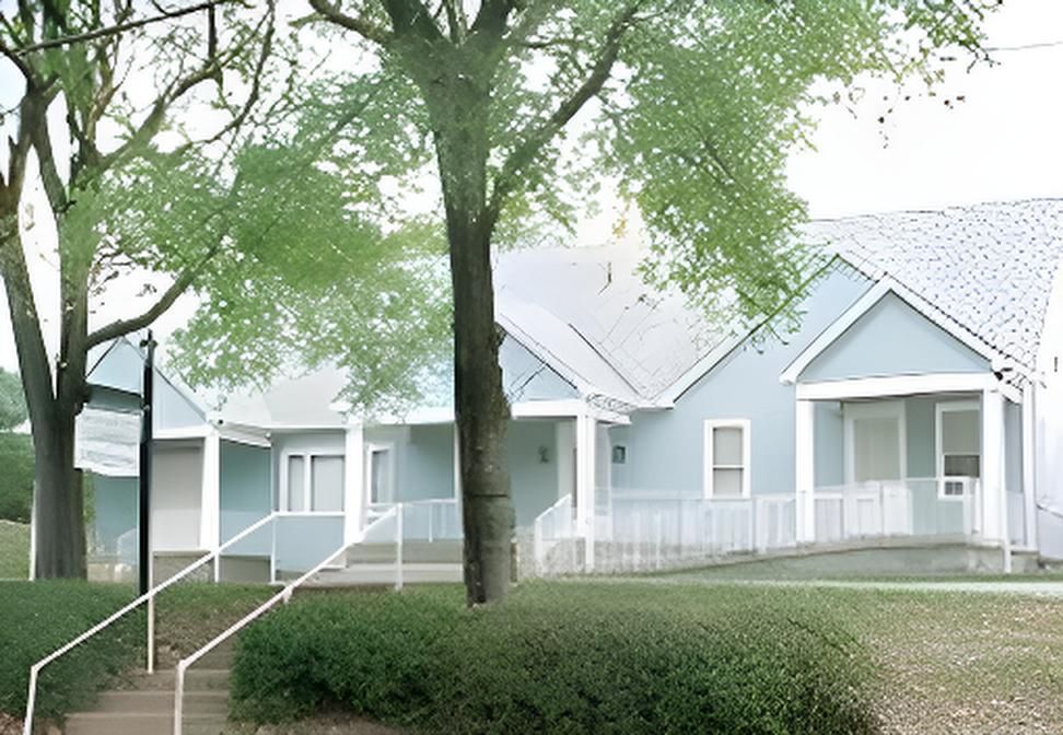 Light blue building with white trim, porch, and a sign, trees in front.