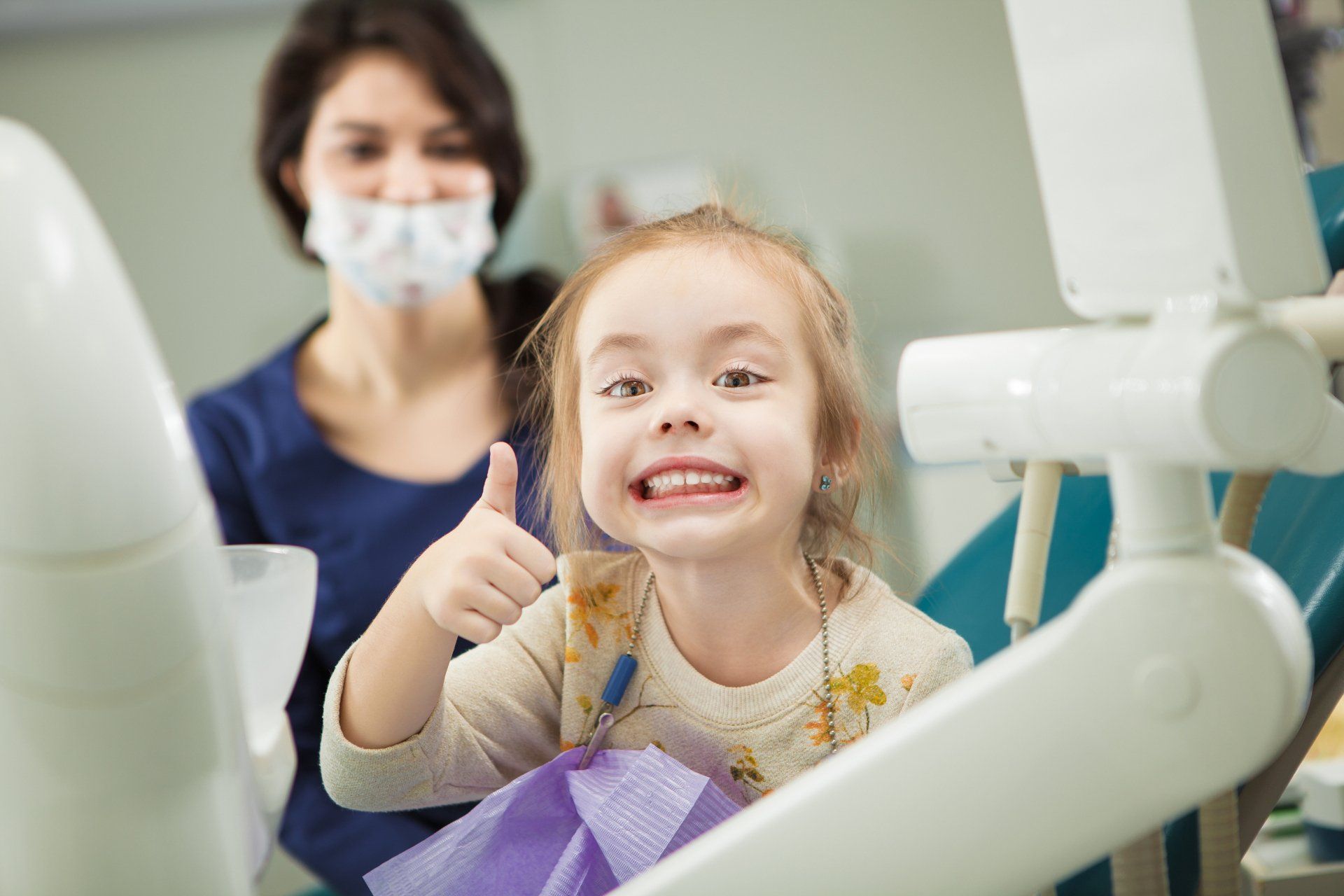 A little girl is sitting in a dental chair and giving a thumbs up.