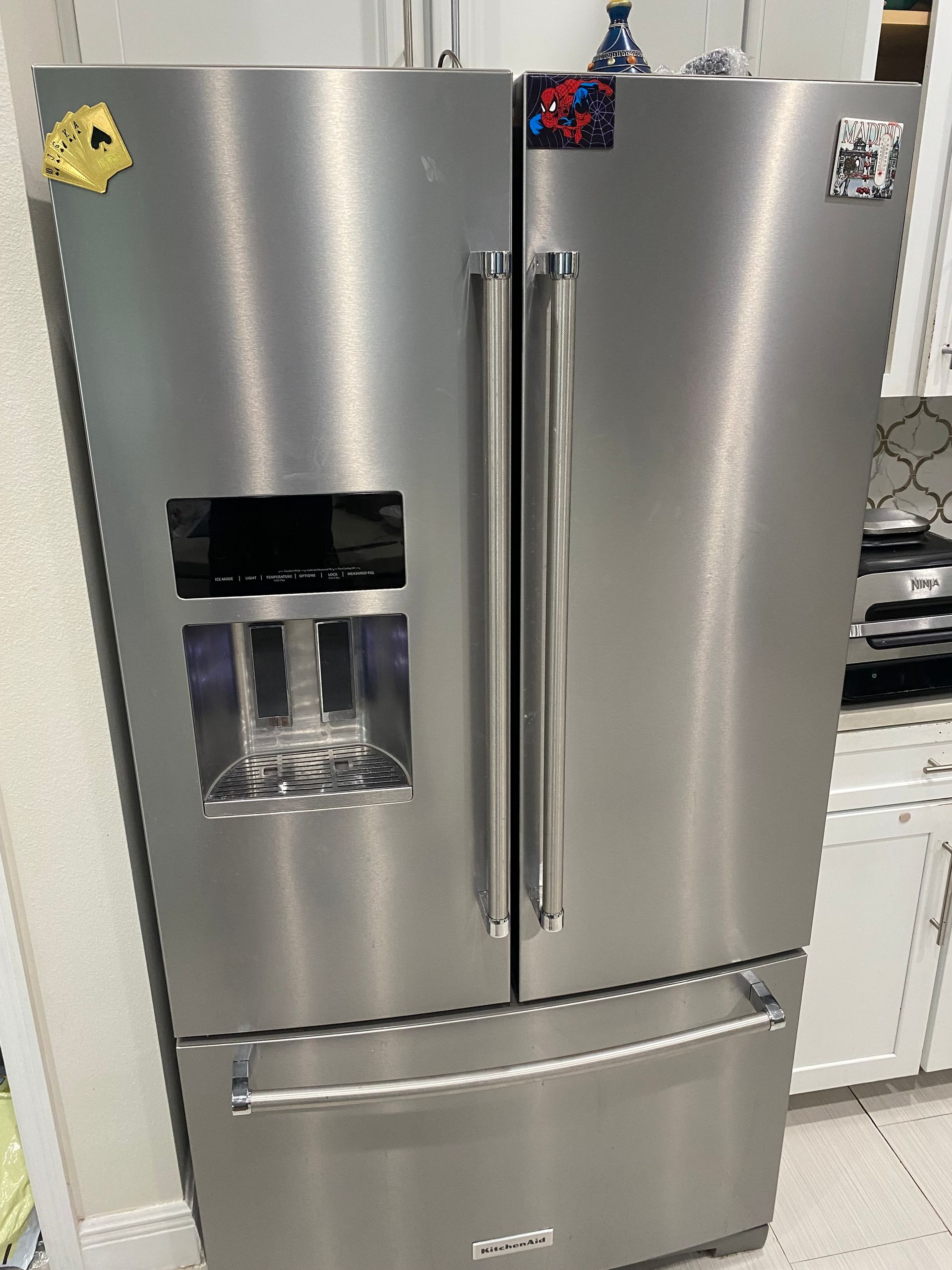 Stainless steel, three-door refrigerator with water dispenser; kitchen setting.