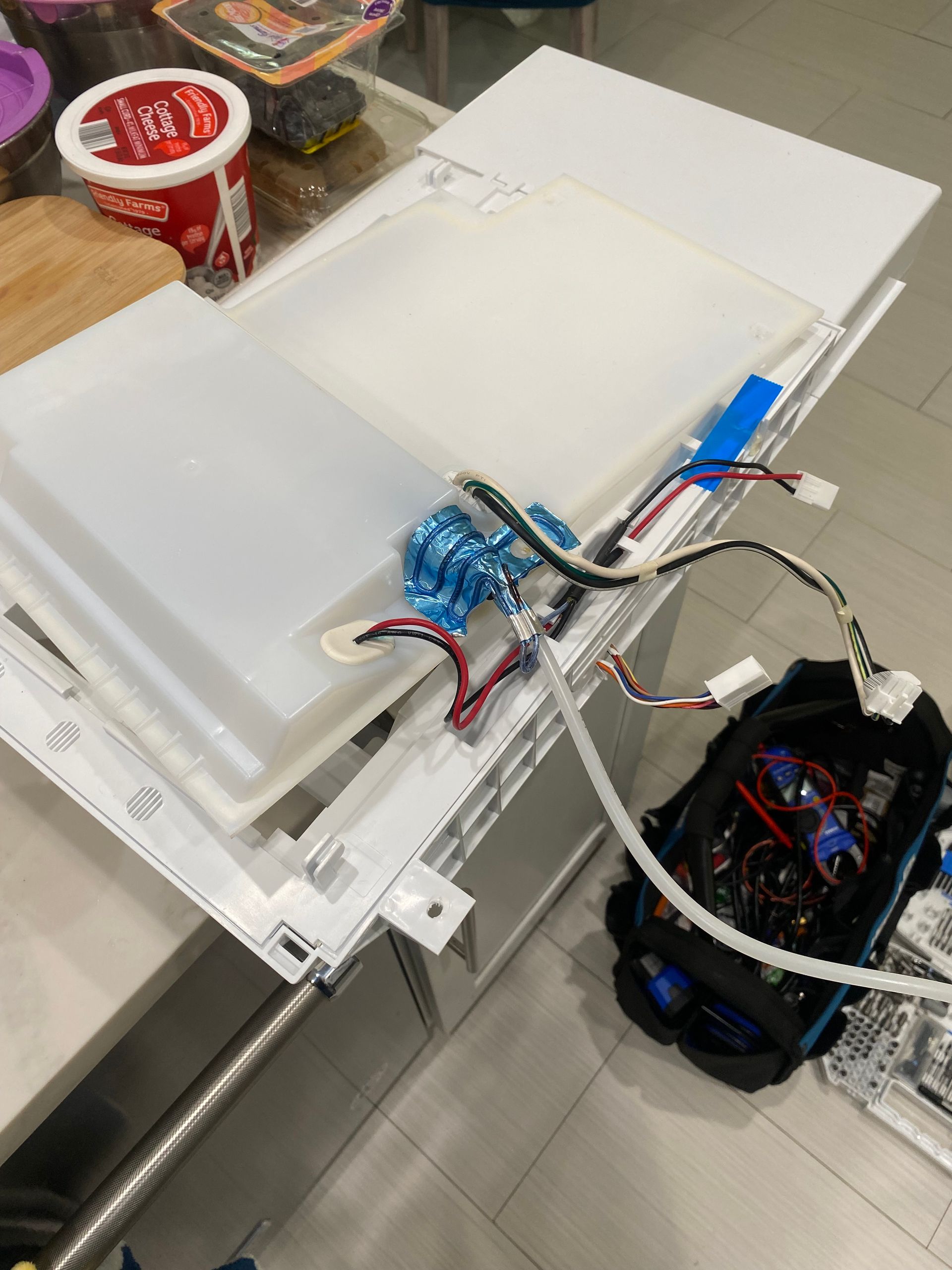 Disassembled refrigerator components on a countertop: white shelves, wiring, and a dark box.