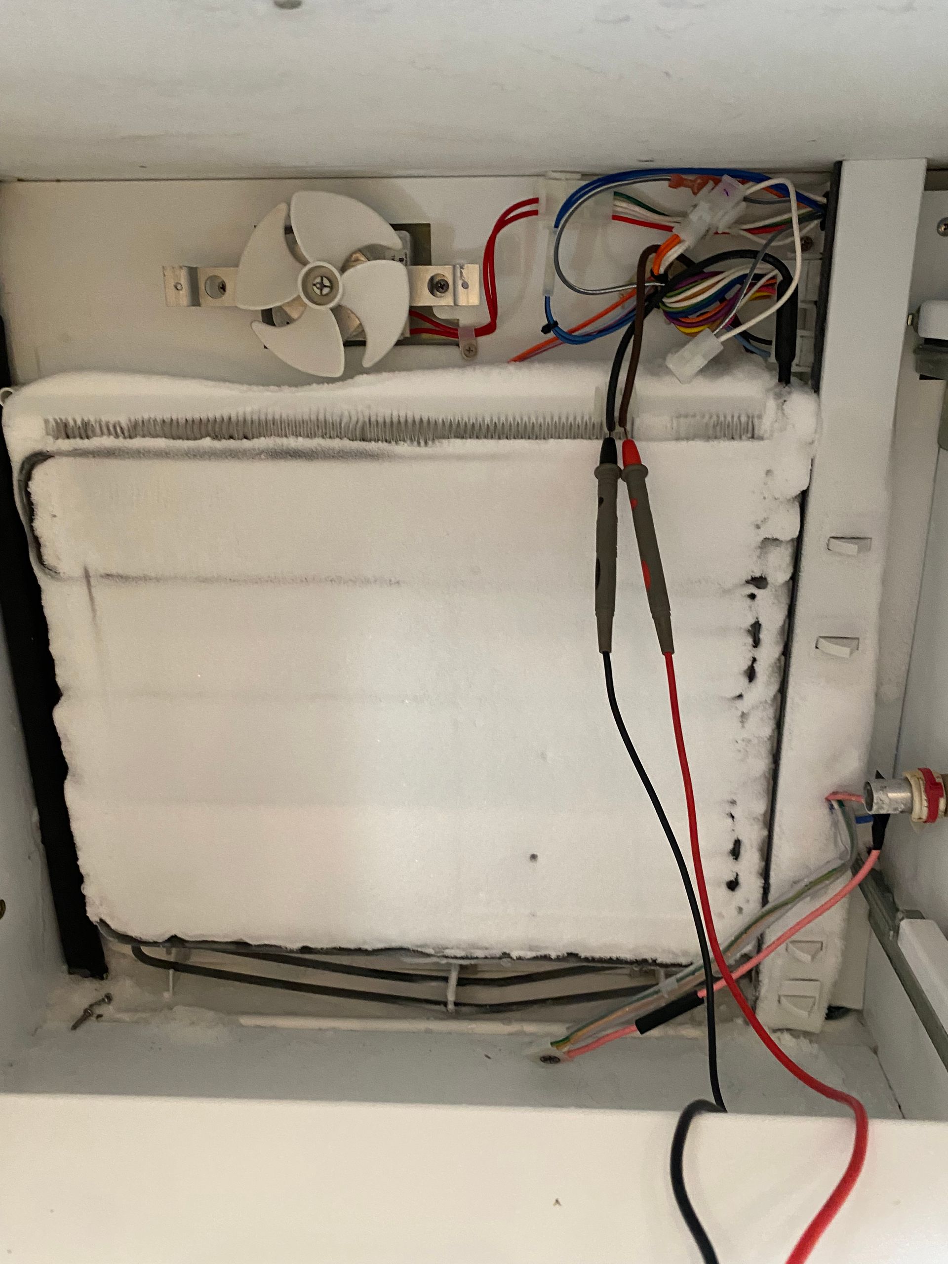 Interior of a freezer heavily frosted with ice, revealing components like a fan, wires, and heating elements.
