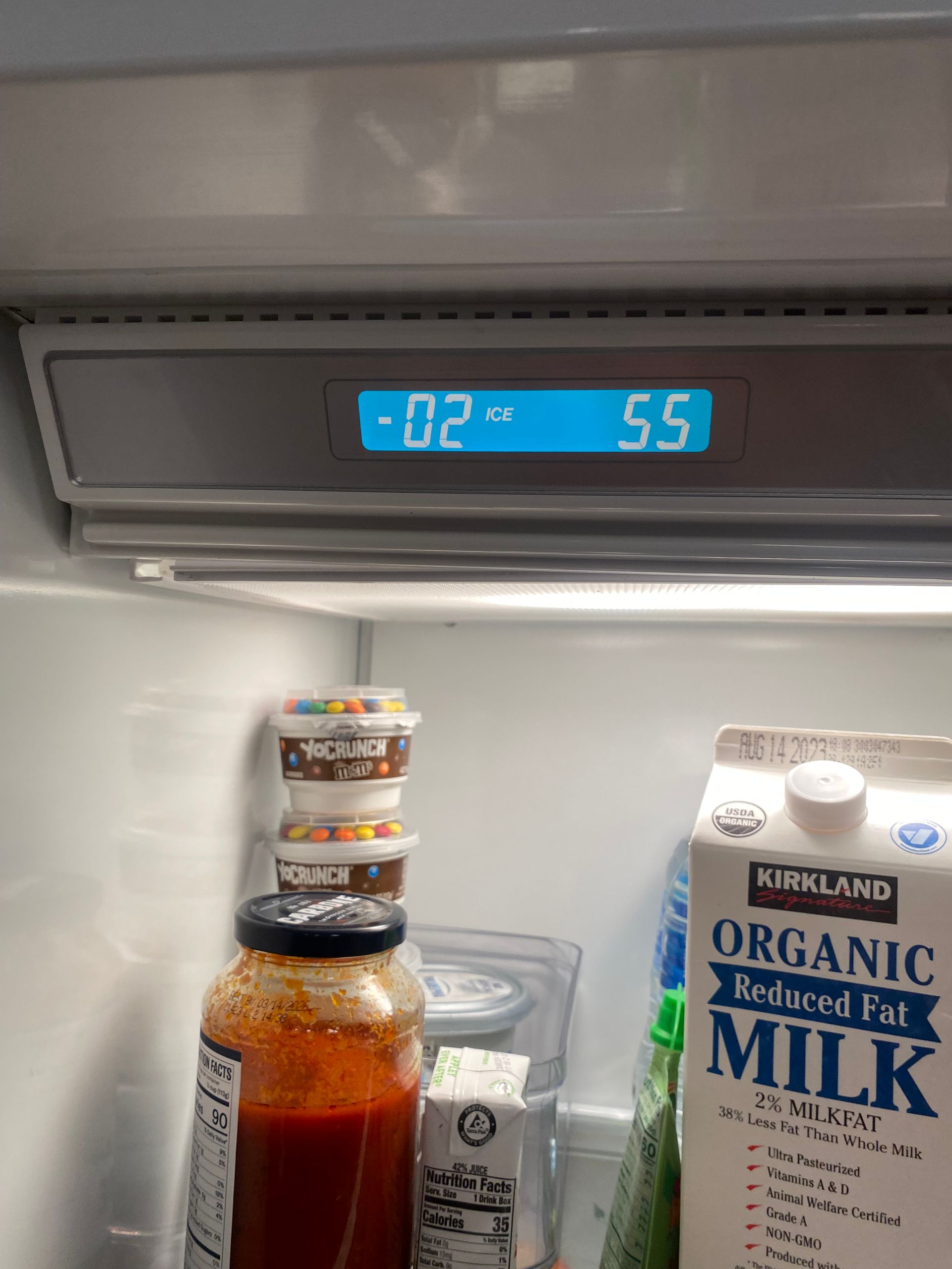 Refrigerator interior with digital display showing -02°C and 5.5. Jars, milk carton, and stacked sweets visible.