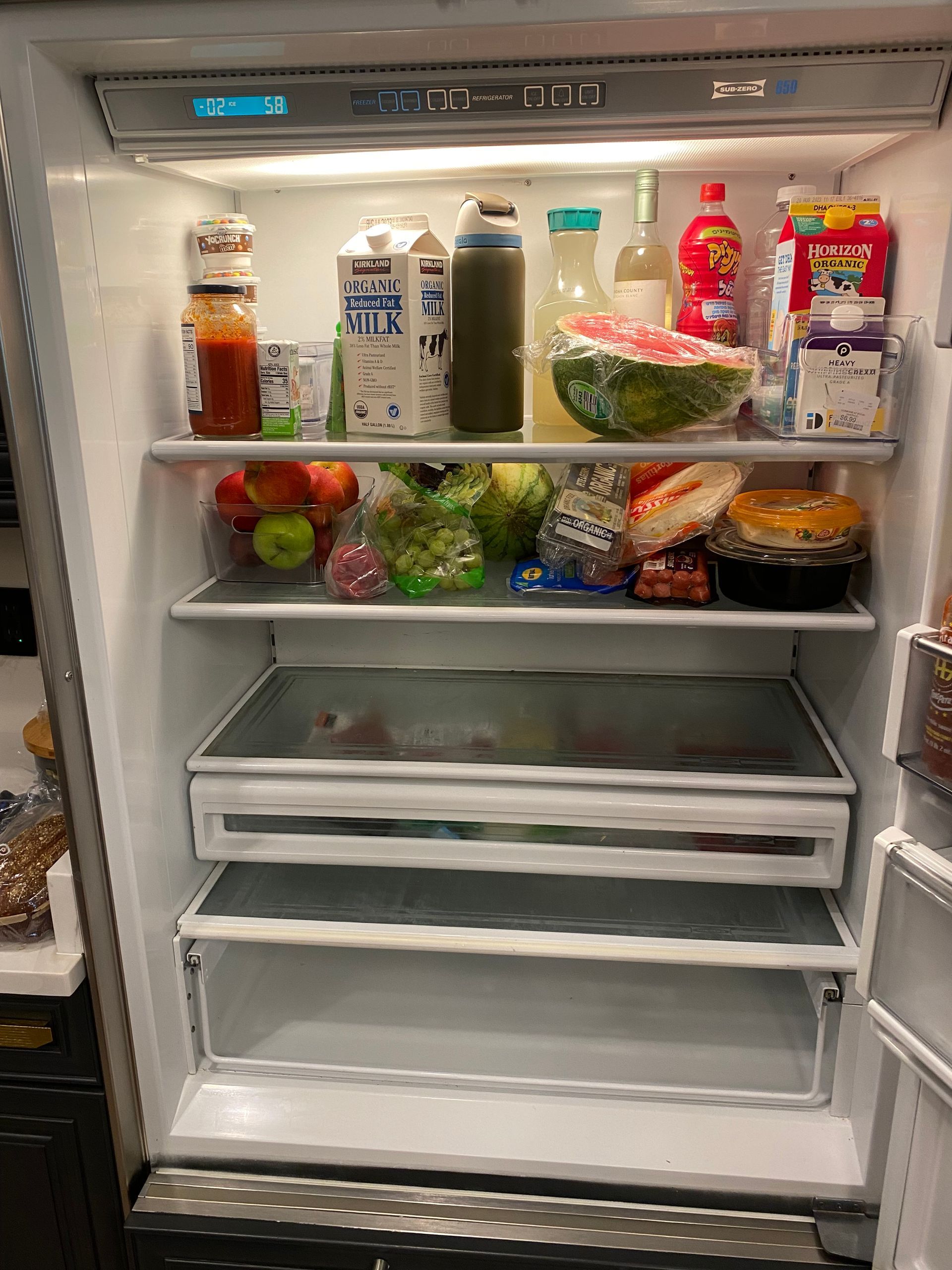 Open refrigerator filled with food: fruits, milk, juice, sauces, and leftovers.