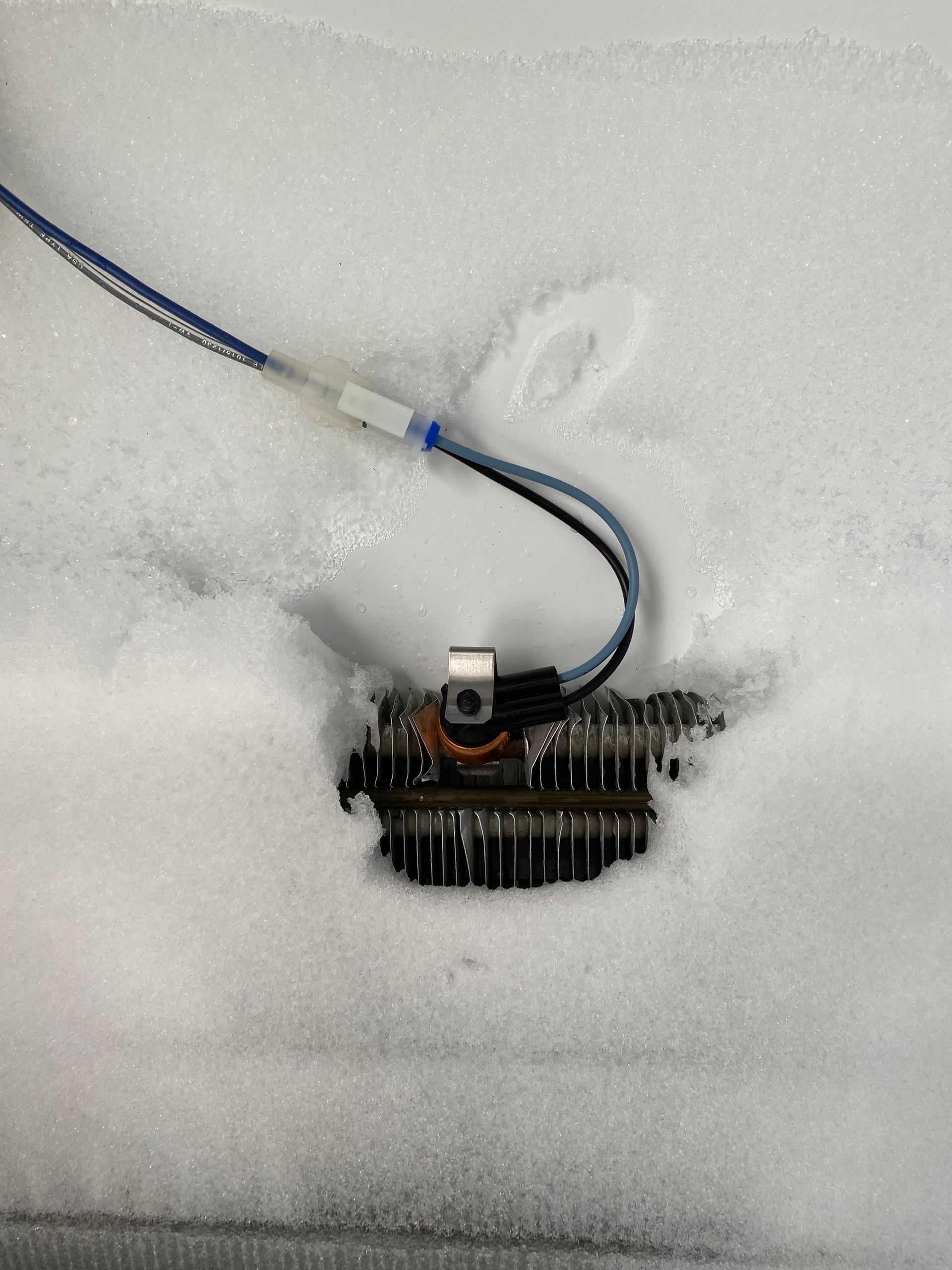 Wires connected to a frozen refrigerator coil. White snow partially covers the coil.