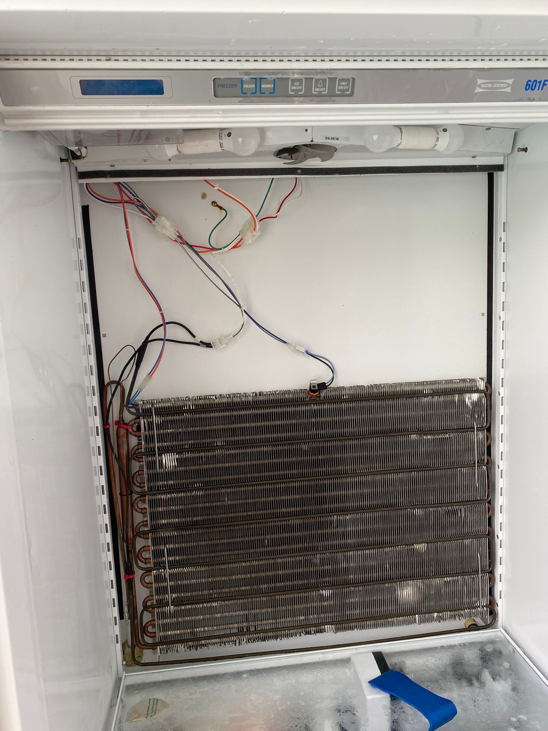 Open refrigerator interior, showing a coil covered in frost and wiring behind a white panel.