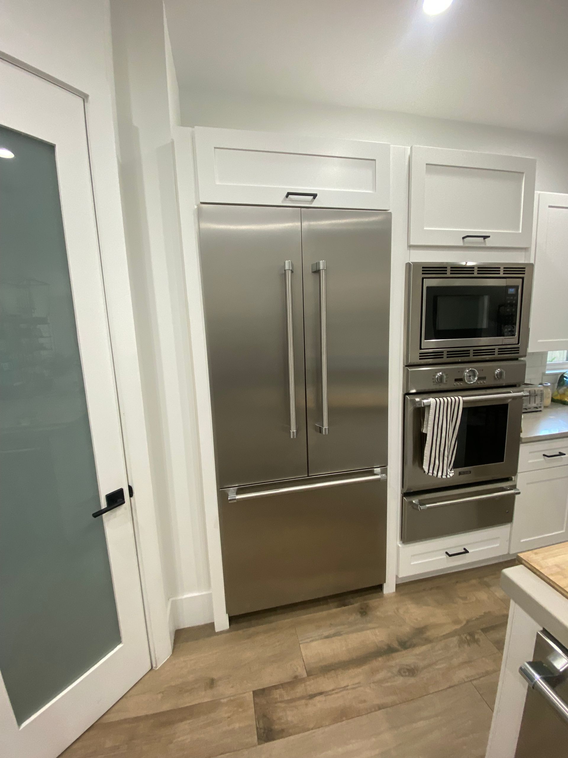 Stainless steel refrigerator and oven built-ins in a modern kitchen. White cabinetry, wood floor.