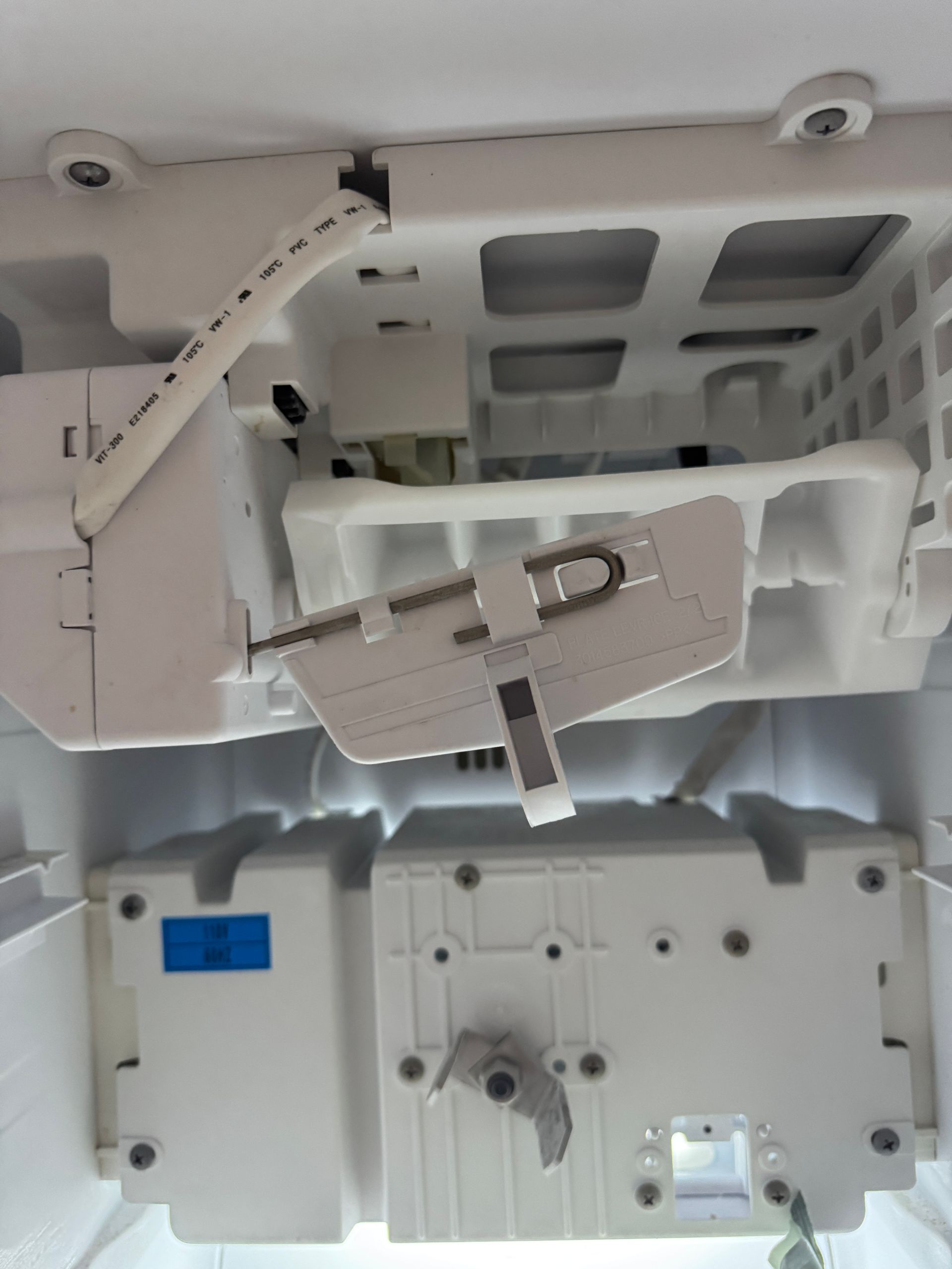 Inside of a refrigerator, ice maker mechanism with a wire arm in white.