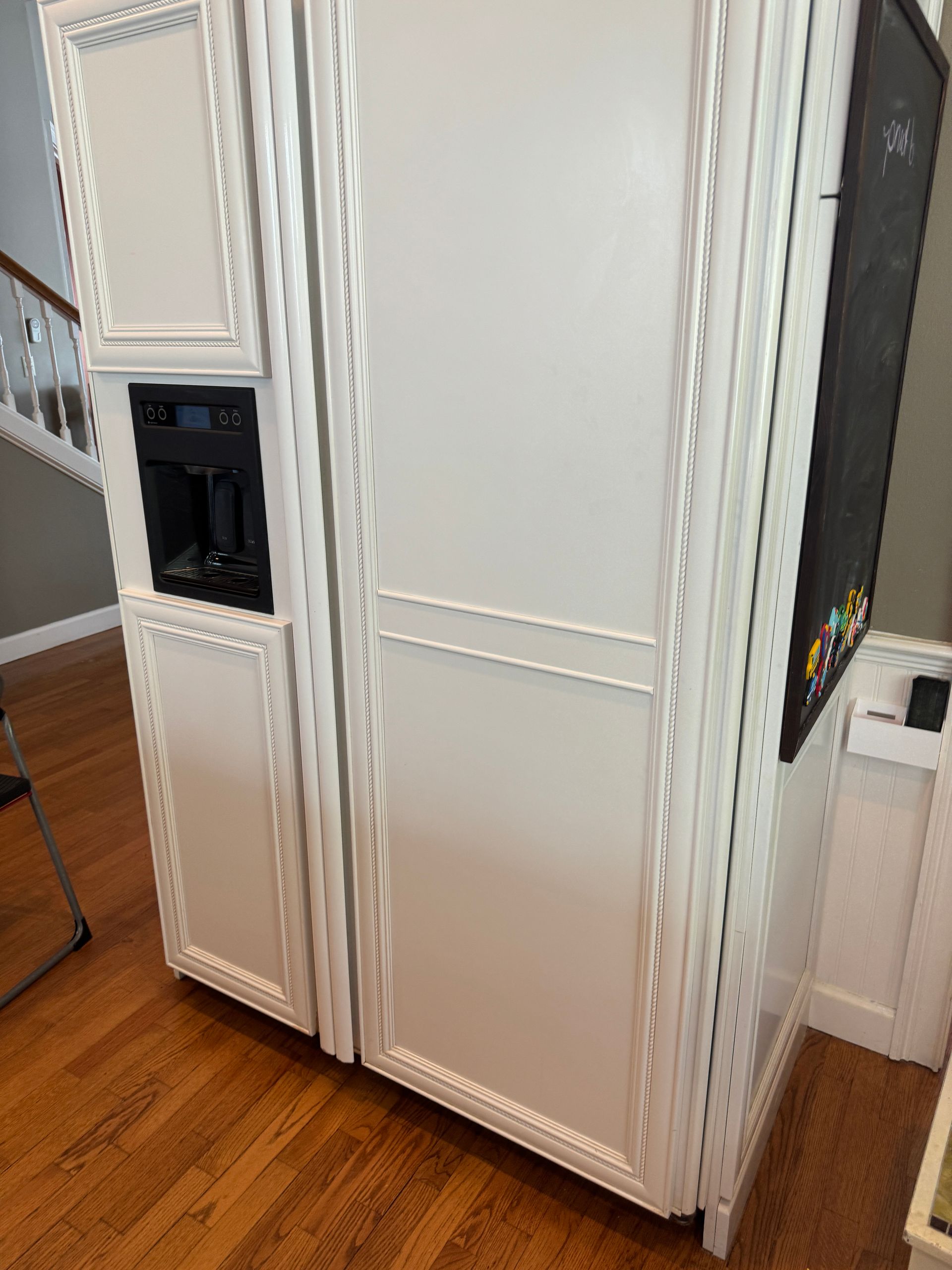 White refrigerator with water dispenser built into white cabinetry; adjacent chalkboard and hardwood floor.